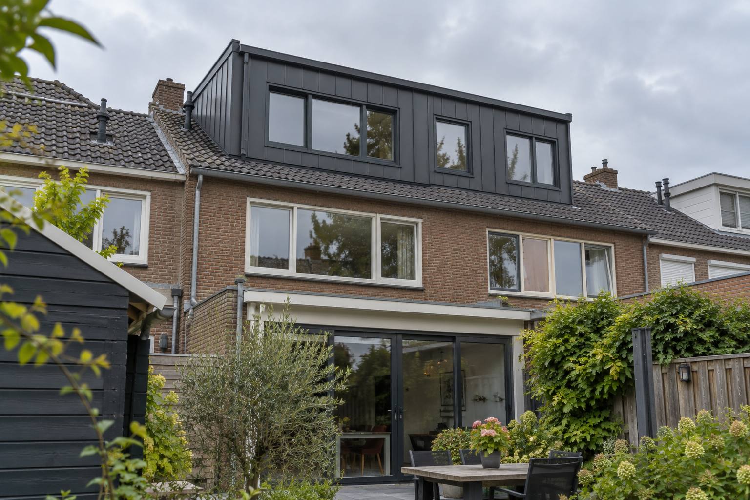 Brede antraciet dakkapel aan de achterkant van een rijwoning, gezien vanuit de tuin